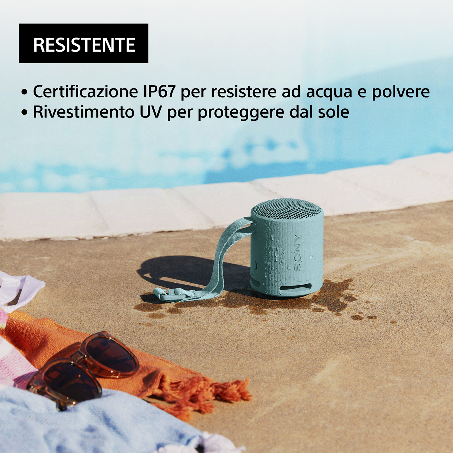 Un piccolo altoparlante turchese, appoggiato su una superficie vicino a una piscina, con occhiali da sole e un asciugamano.