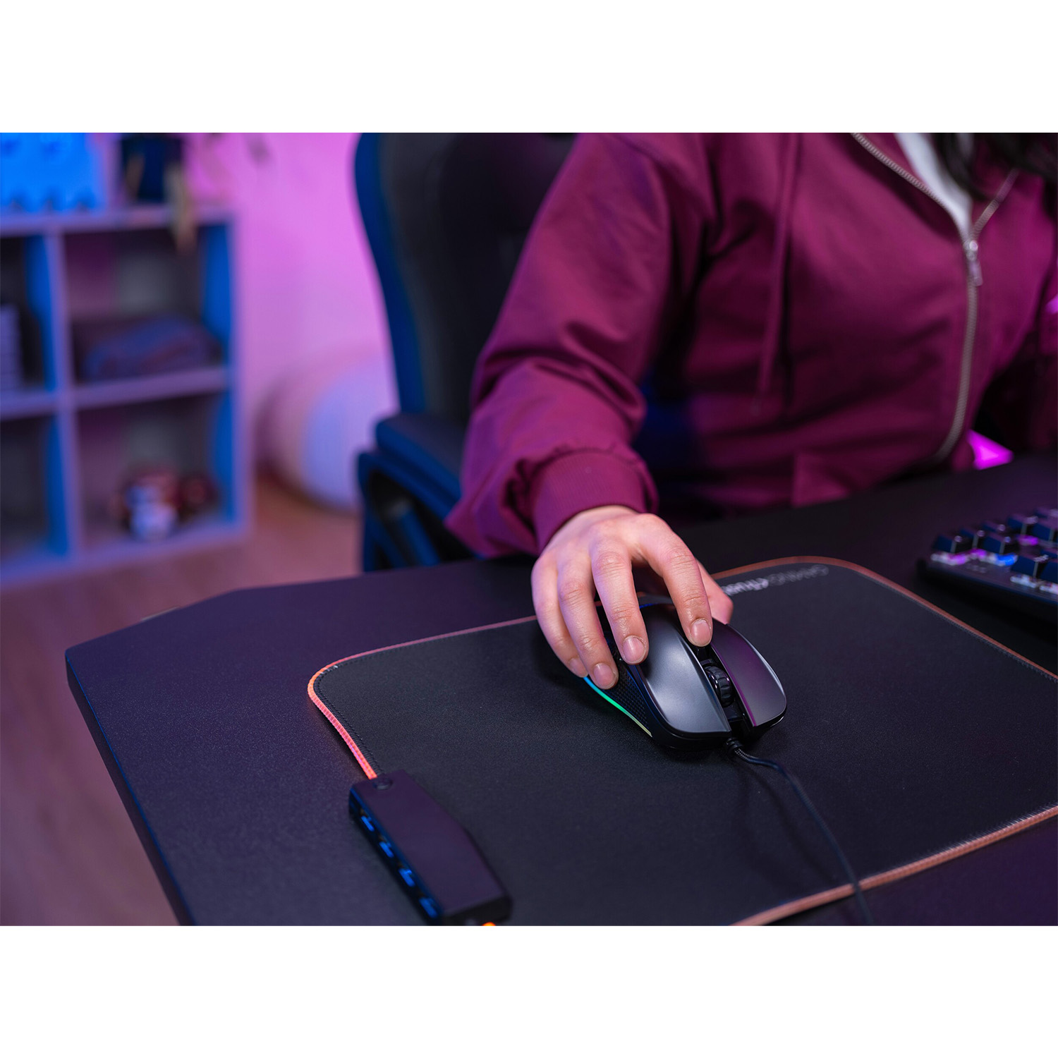 Mano che usa un mouse da gioco nero su un tappetino. Scrivania scura, luci viola e blu.
