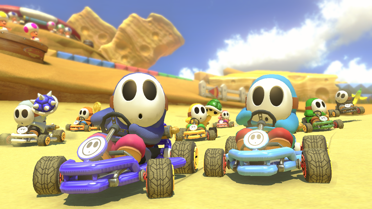 Characters in kart racing cars on a sandy track under a blue sky. The characters are Shy Guys.