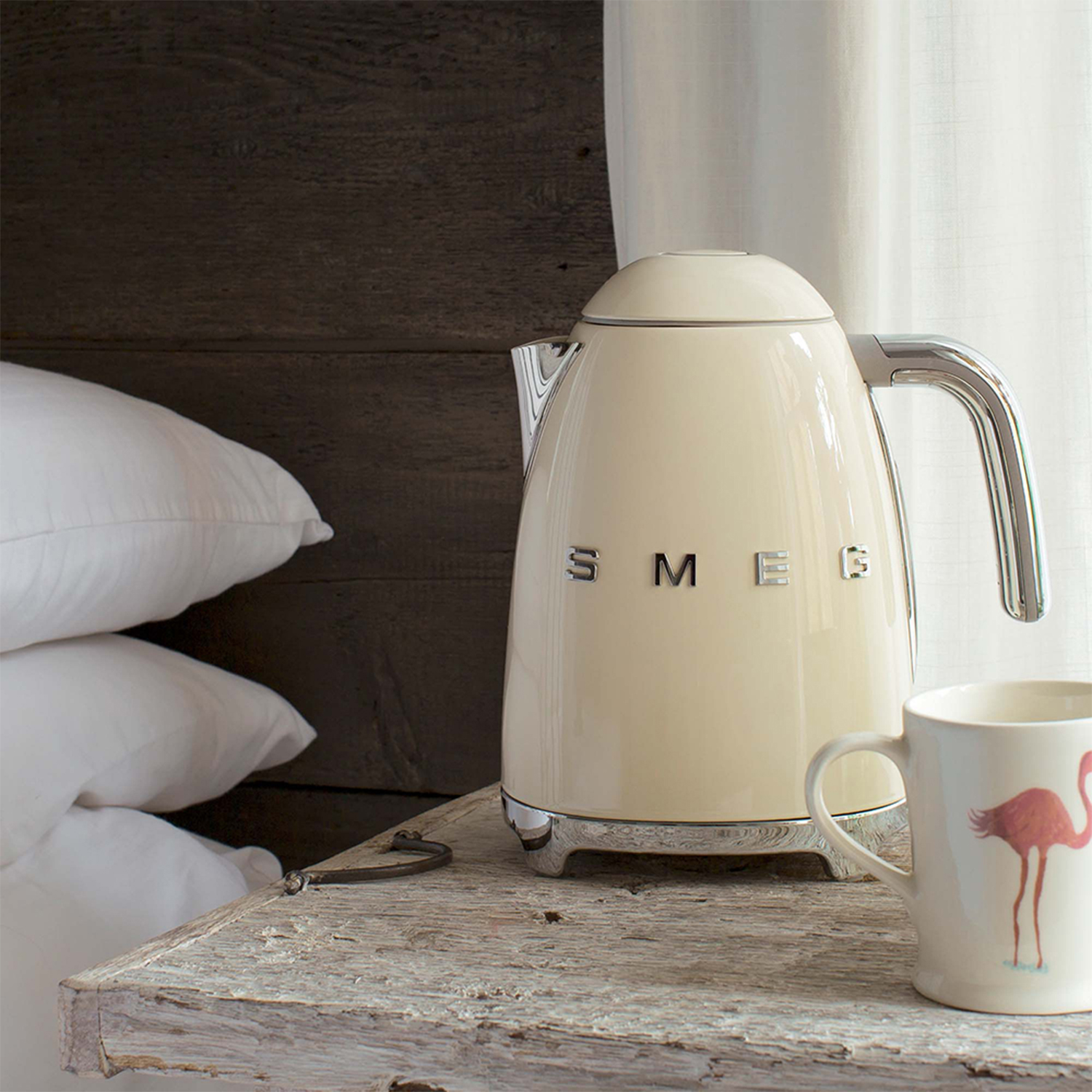 Bollitore elettrico beige su una superficie di legno con una tazza e cuscini sullo sfondo.