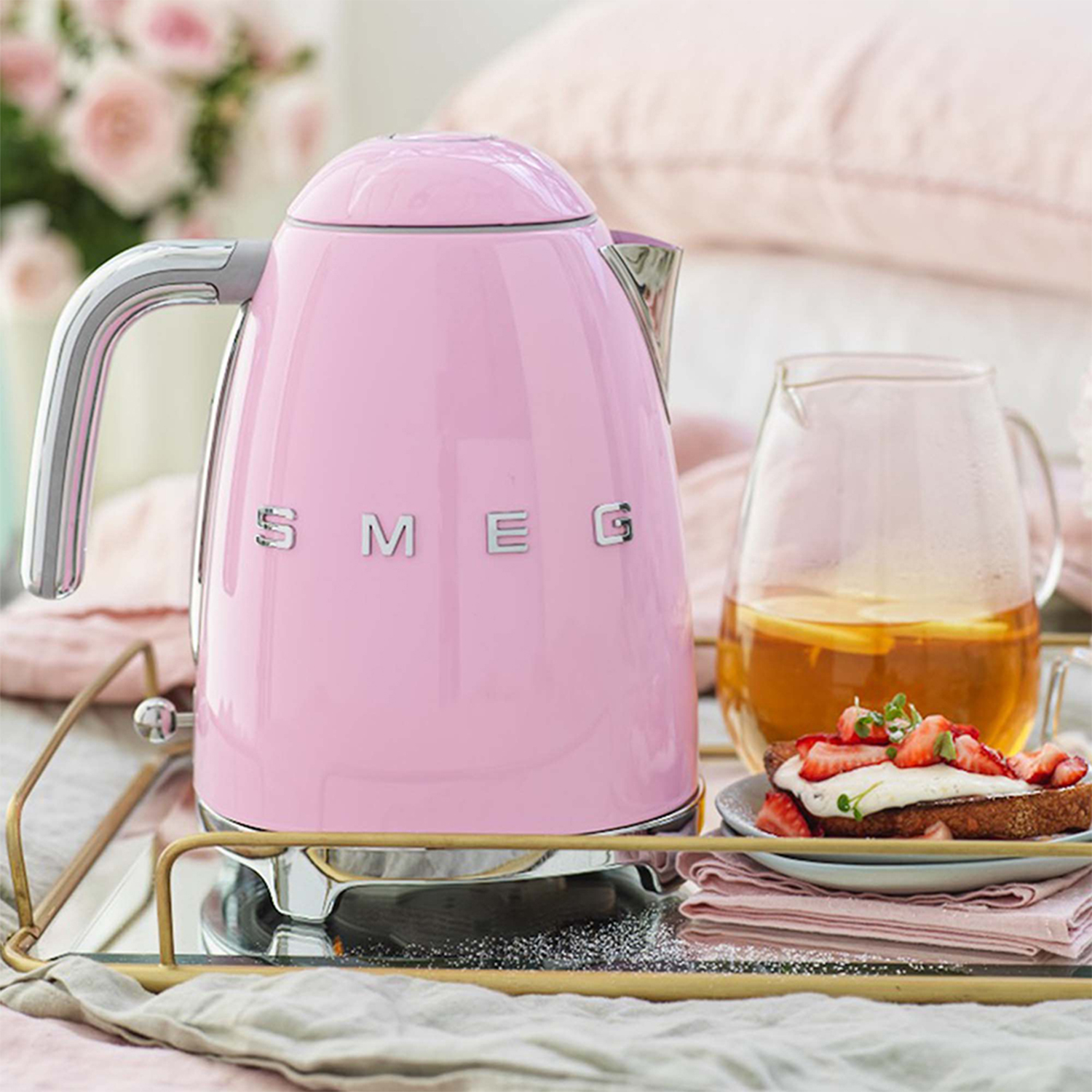 Bollitore SMEG rosa su un vassoio a specchio con toast e fragole, cuscini rosa sullo sfondo.