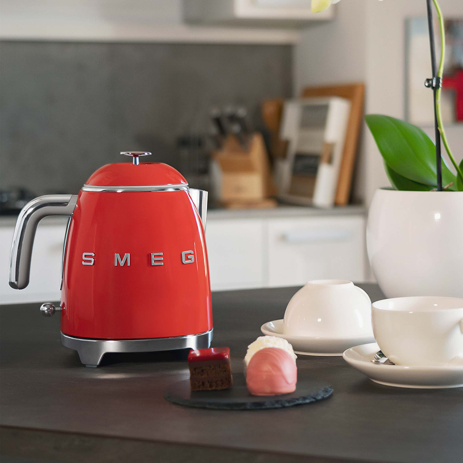 Bollitore Smeg rosso, piccole torte e tazze sul bancone. Sfondo cucina bianco.