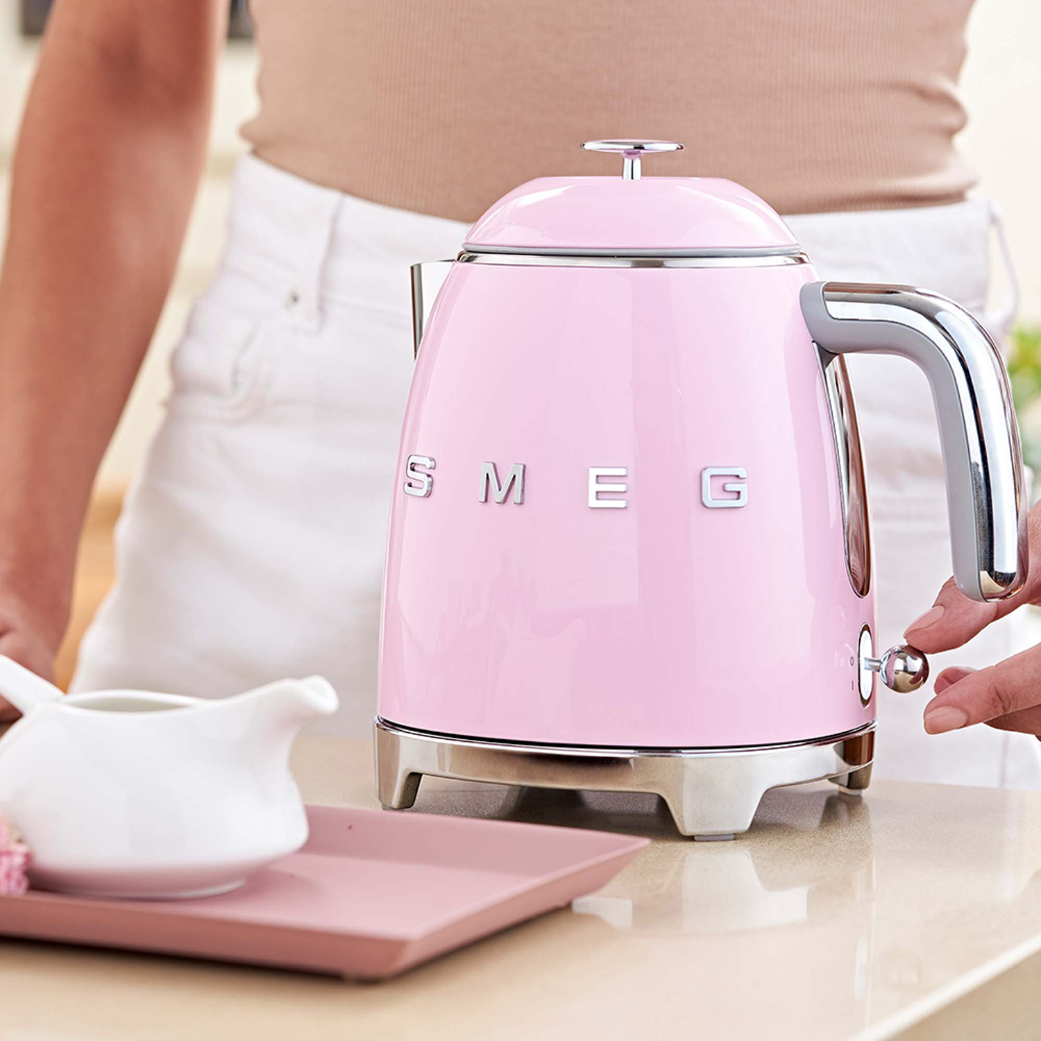 Bollitore Smeg rosa su un vassoio. Mano ruota il quadrante. Sfondo bianco.