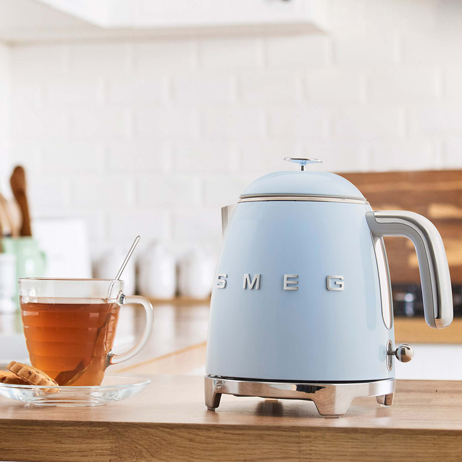 Bollitore Smeg blu su superficie di legno, con tazza da tè e biscotti.