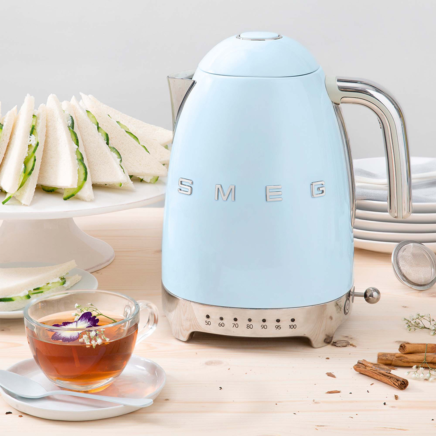 Un bollitore Smeg blu accanto a una tazza di tè, tramezzini e altri oggetti su una superficie di legno chiaro.