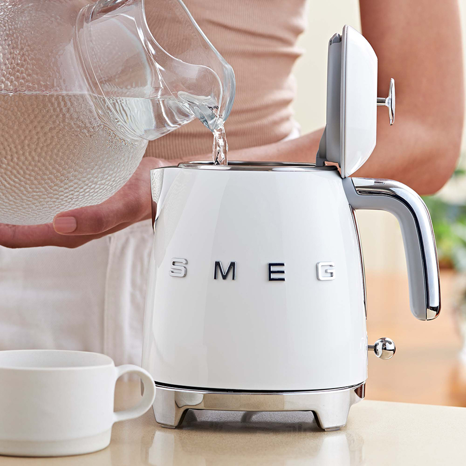Un bollitore Smeg bianco che viene riempito d'acqua da una brocca. Una tazza bianca è sul tavolo.