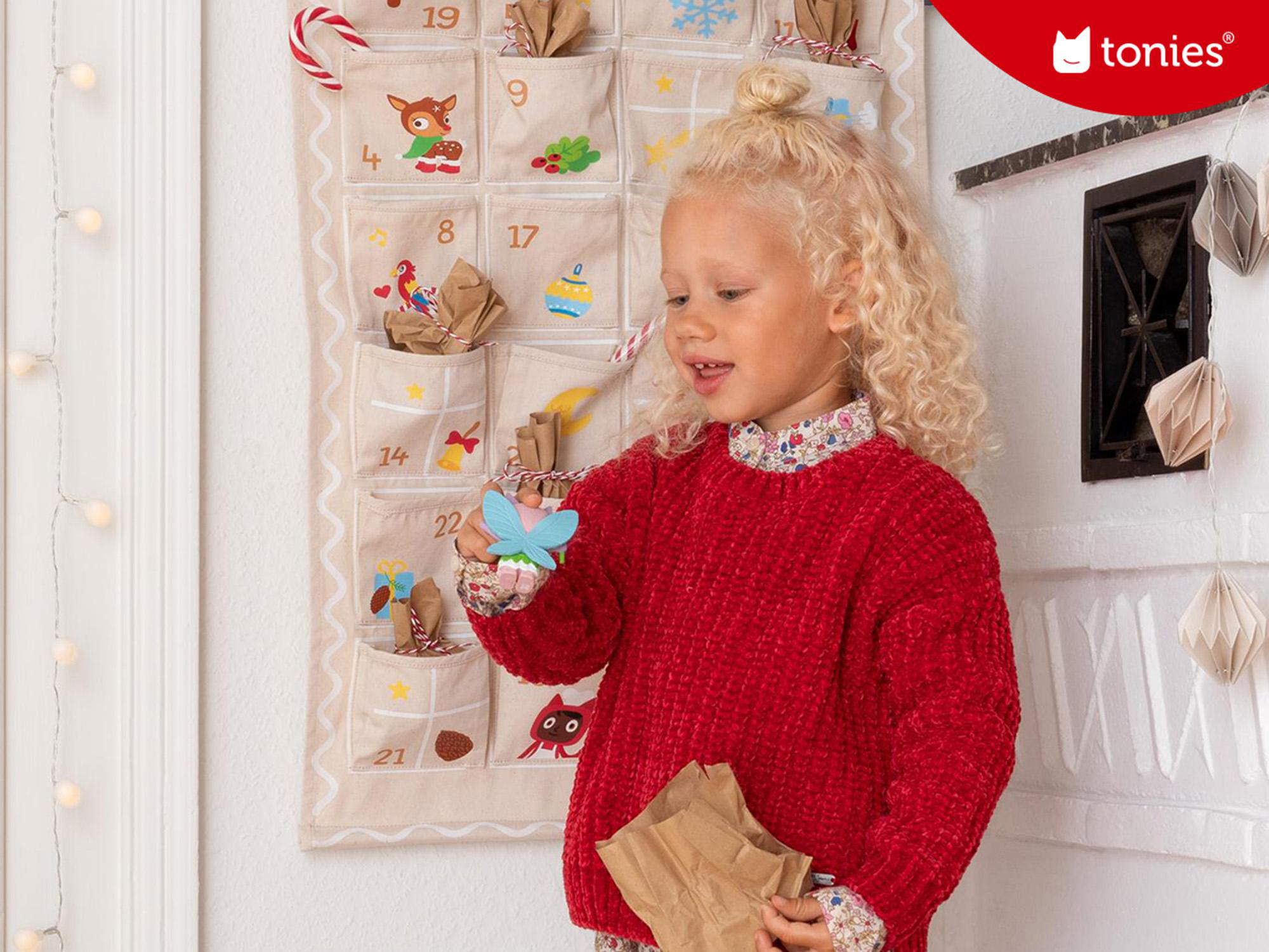 Ein kleines Mädchen in einem roten Pullover öffnet eine Papiertüte neben einem Adventskalender.