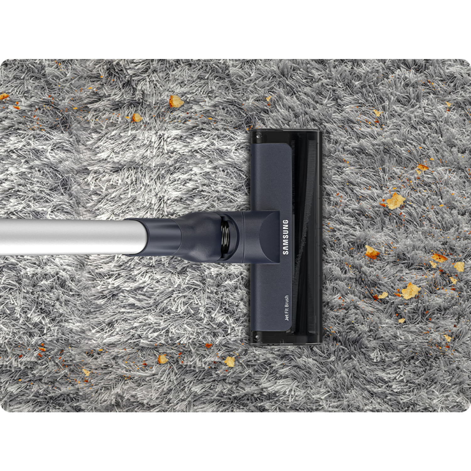 Vacuum cleaner head on a shaggy carpet with scattered debris. Shows Samsung branding. Grey and brown tones.