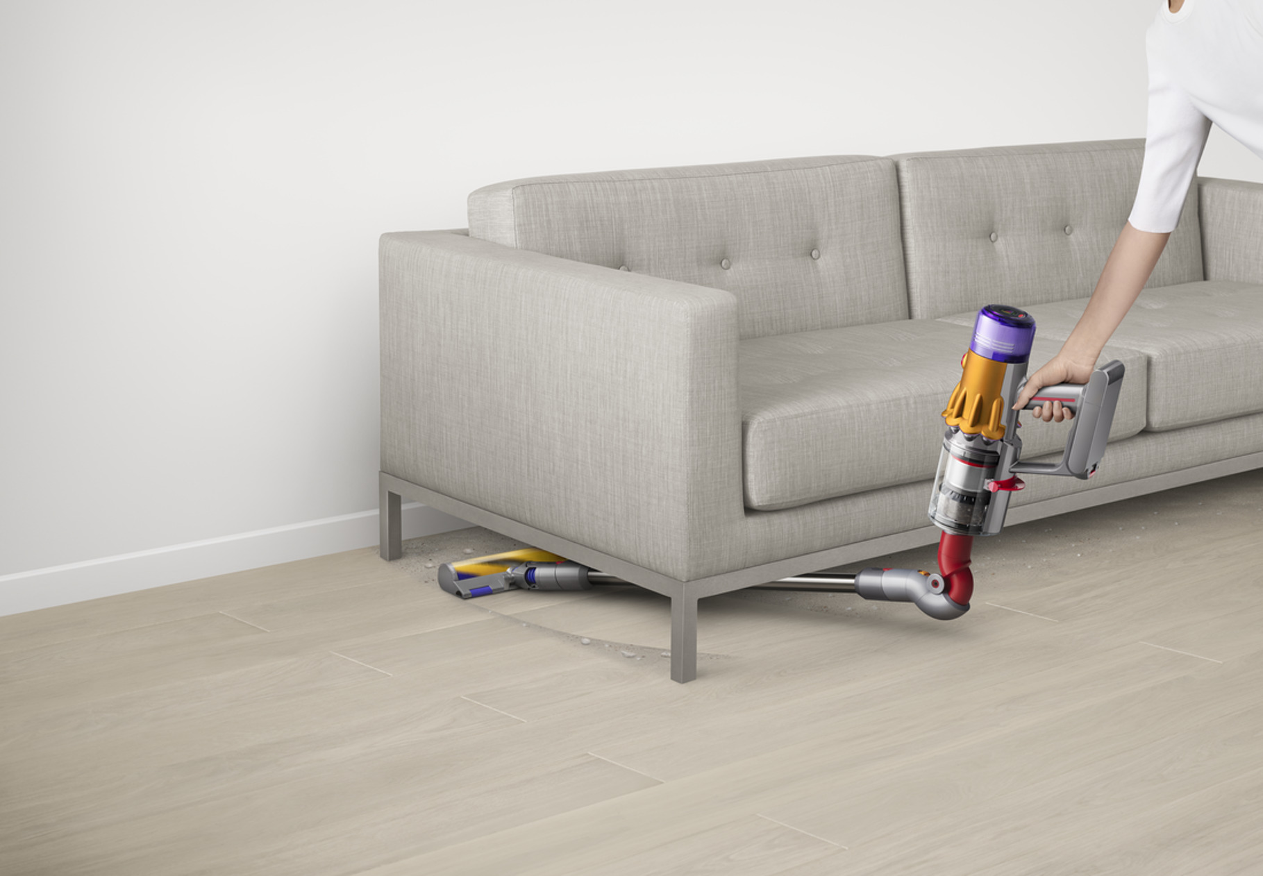 Una persona aspirando debajo de un sofá gris con una aspiradora Dyson. Piso de madera clara.