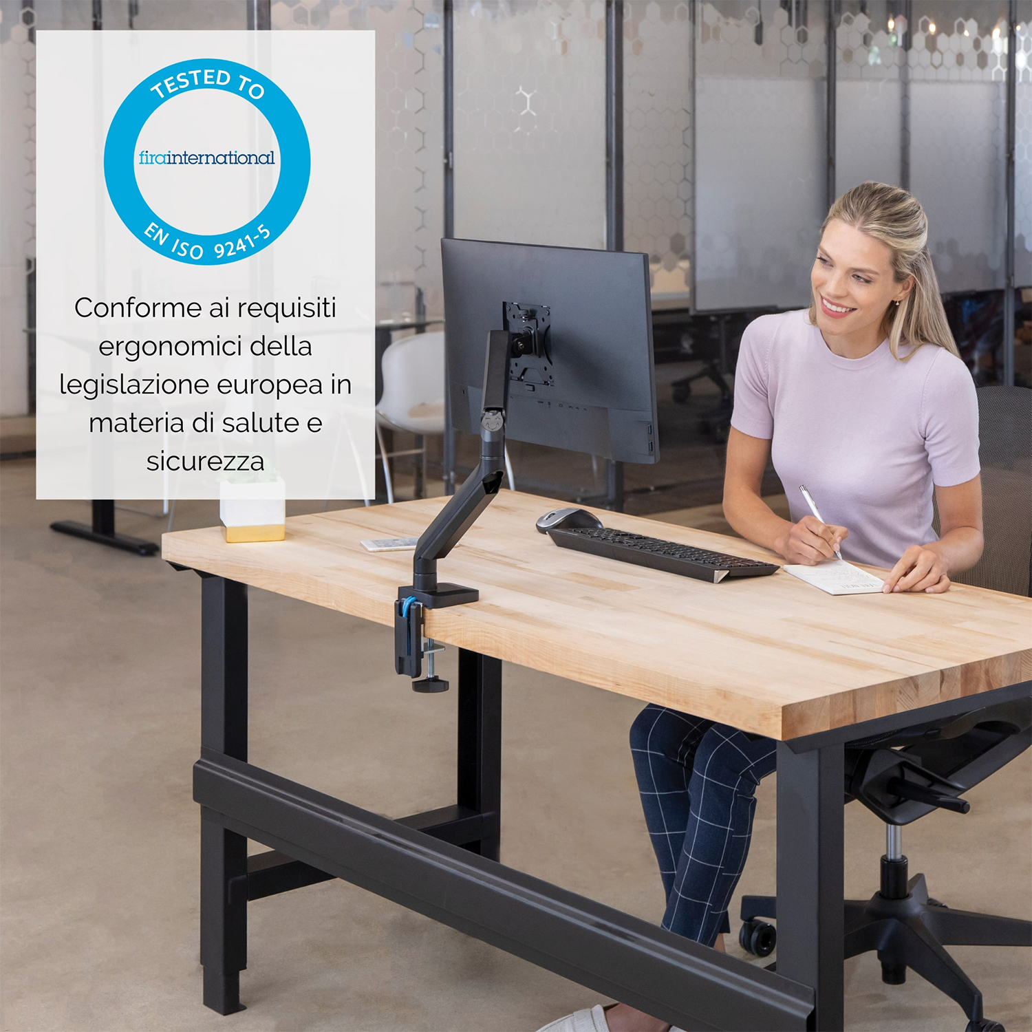 Donna che scrive a una scrivania con supporto monitor. Cartello 'Tested to EN ISO 9241-5'.