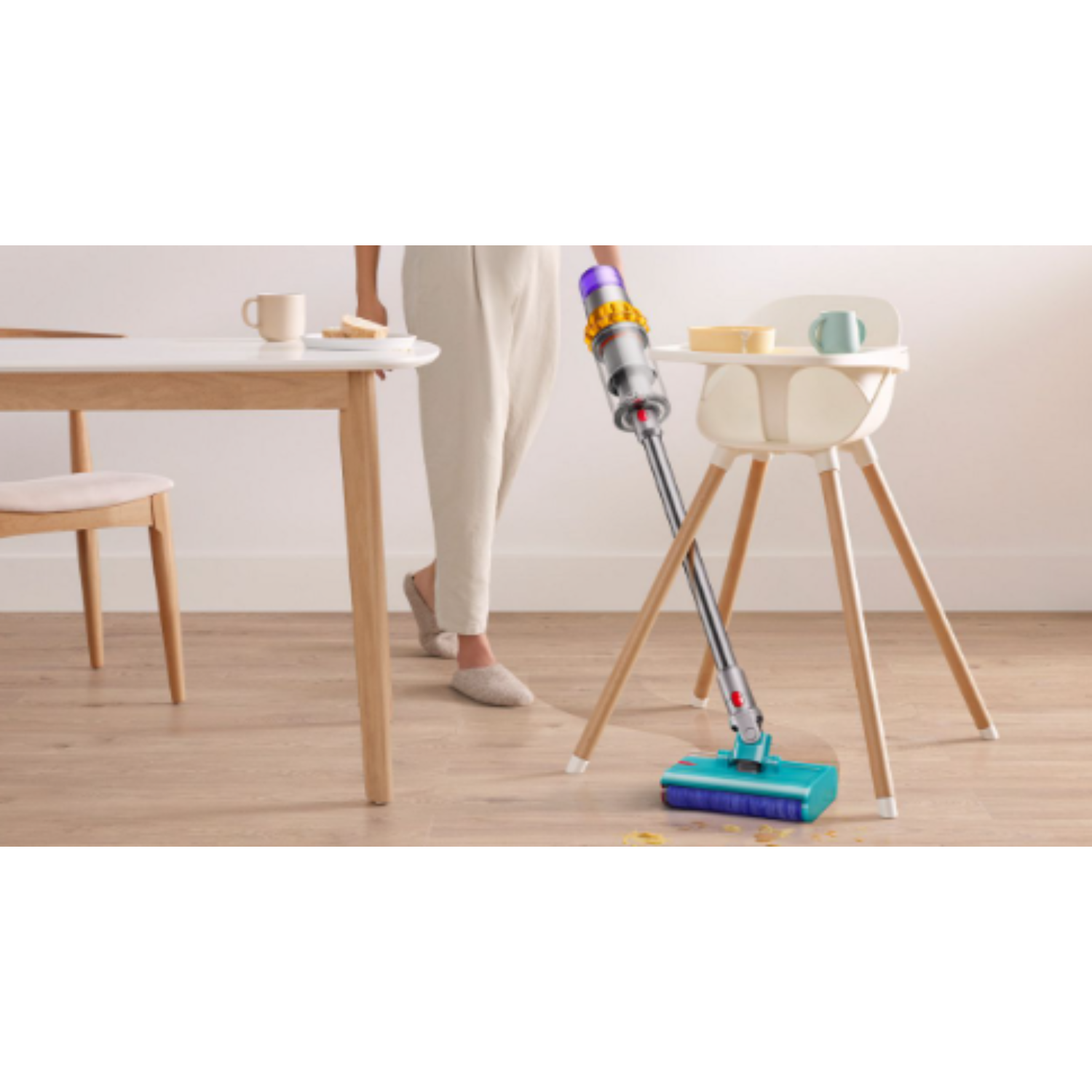 Person vacuuming a wooden floor; high chair and table in the background.