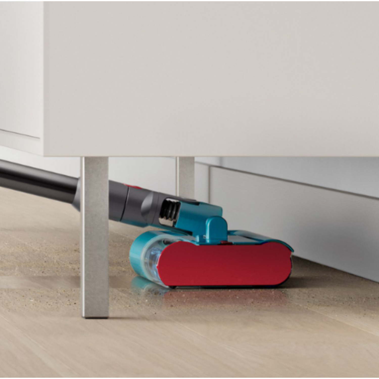 A vacuum cleaner head cleaning under a cabinet, with a teal and red color scheme.