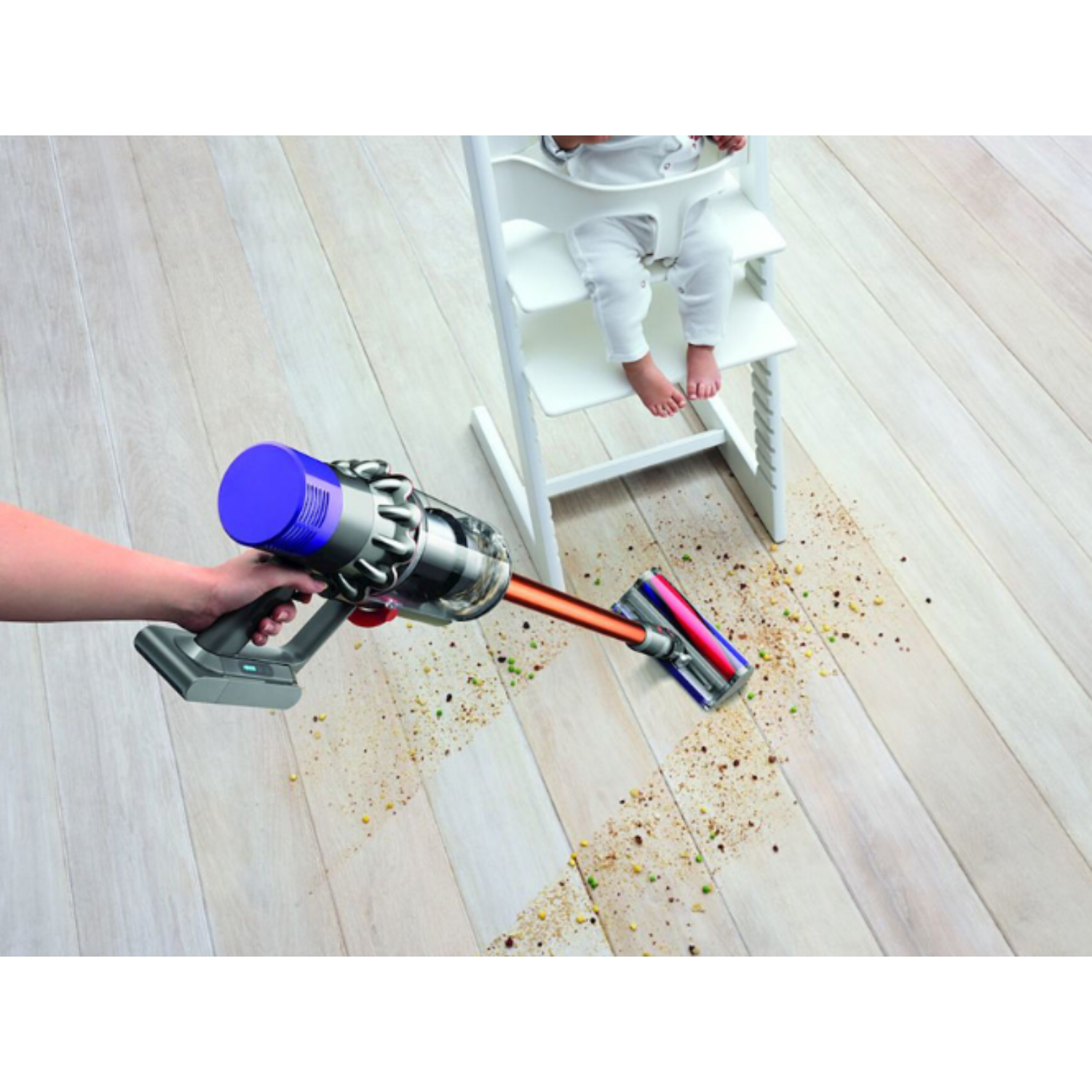 A hand holds a vacuum, cleaning a floor with scattered food debris. A child sits in a high chair.