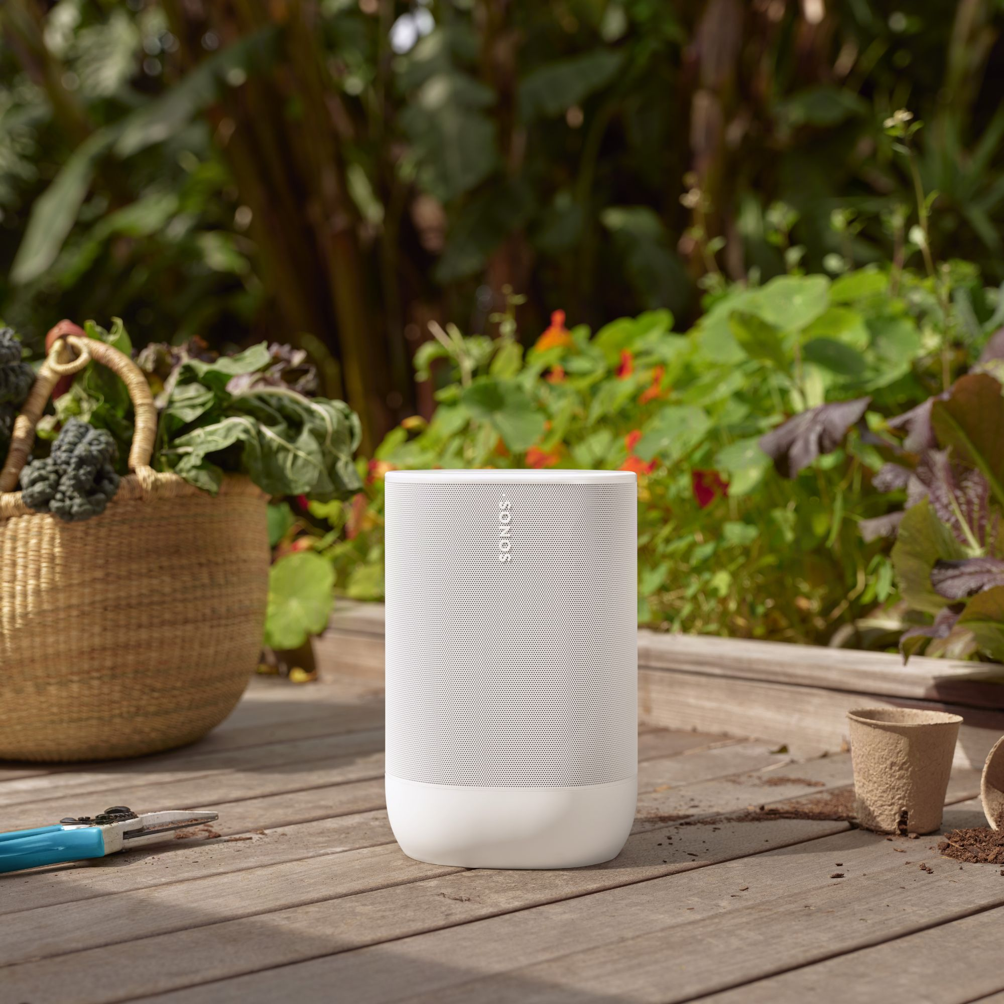 Un altavoz blanco junto a una cesta de verduras y herramientas de jardinería.