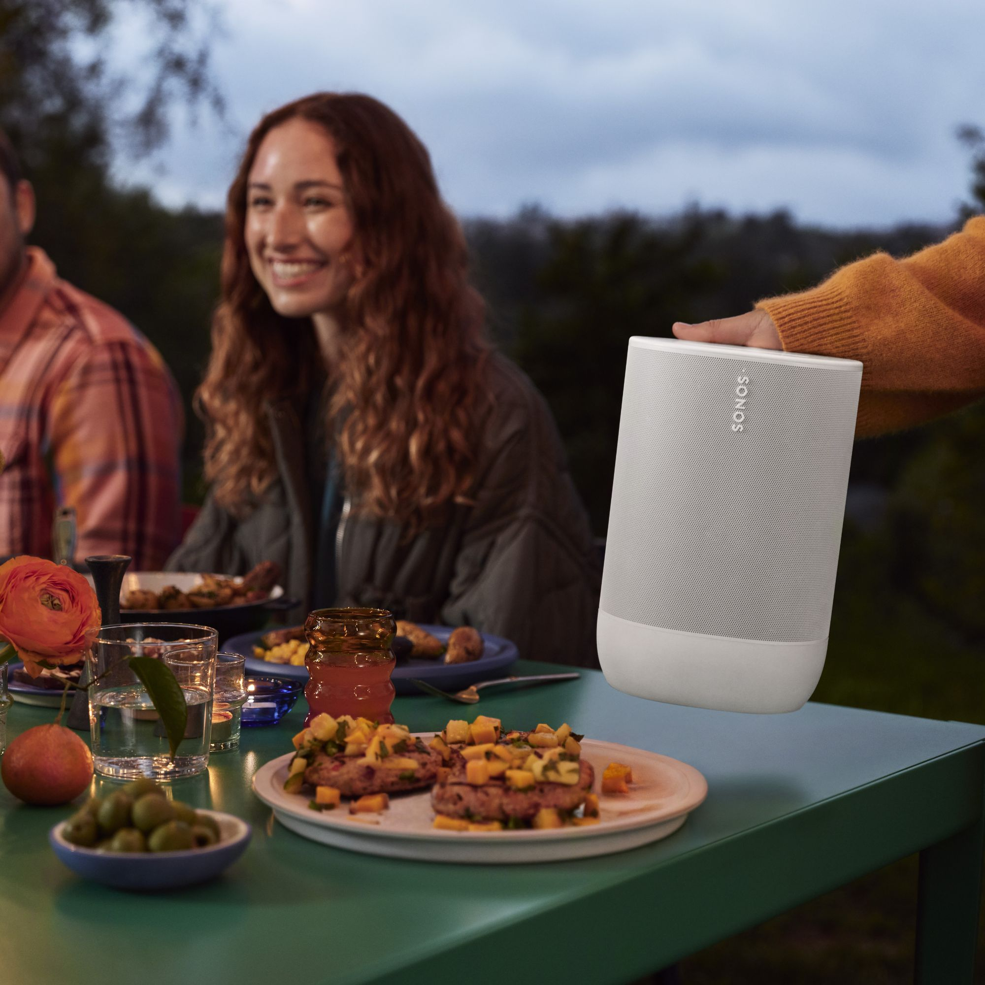 Un altavoz sostenido por una persona, con un grupo de personas al fondo. Mesa con comida y bebidas.