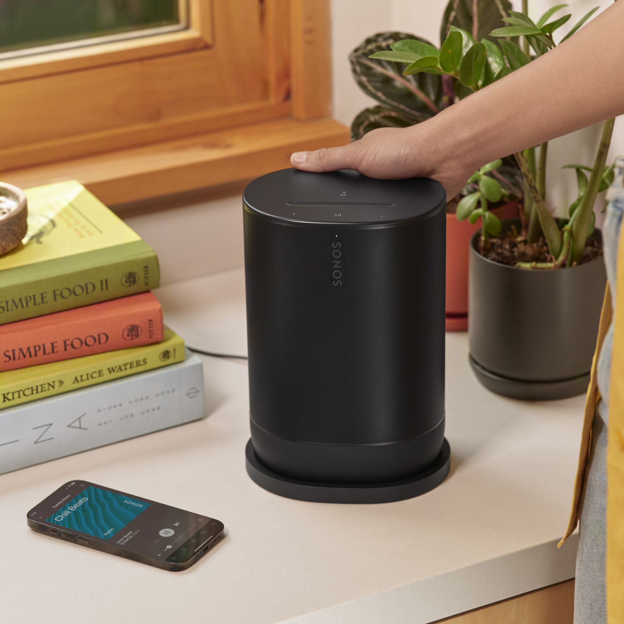 Un altavoz negro está en una encimera, una mano lo toca. Un teléfono con una aplicación de música y libros están cerca.
