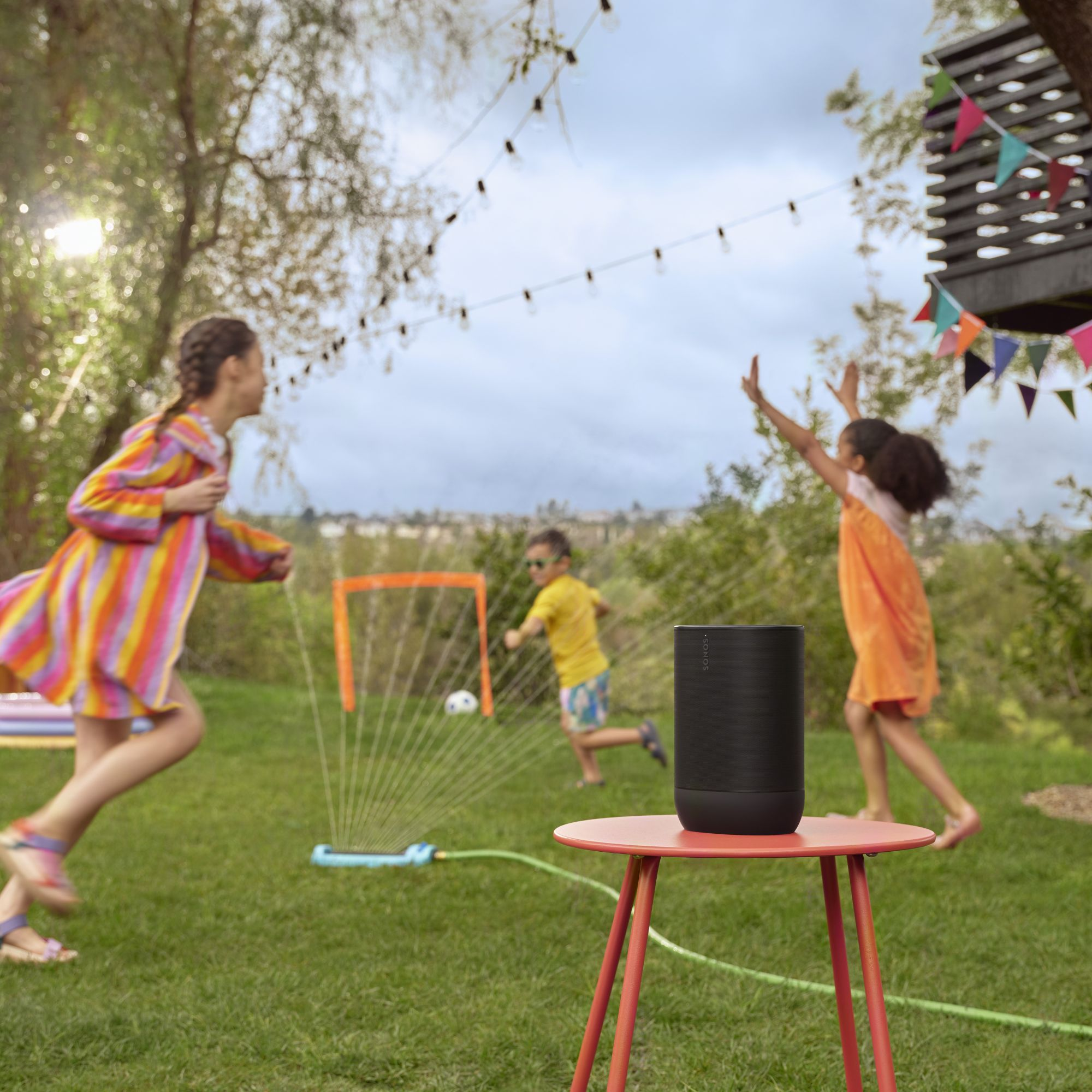 Niños jugando en un jardín con un altavoz en una mesa. Hay un juego de fútbol en curso. El cielo está nublado.