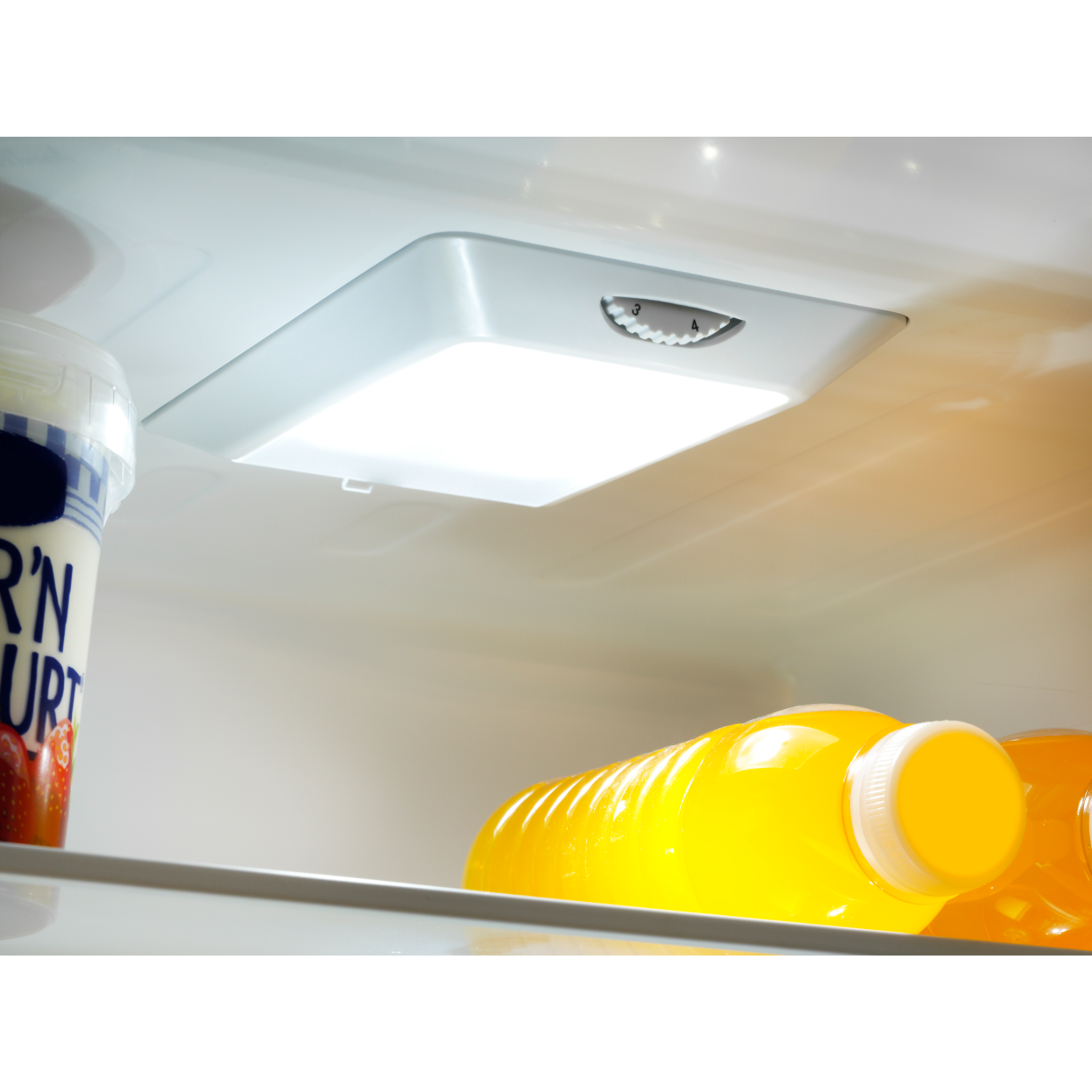 Inside a refrigerator, a light shines on a container of yogurt and bottles of juice on a shelf.