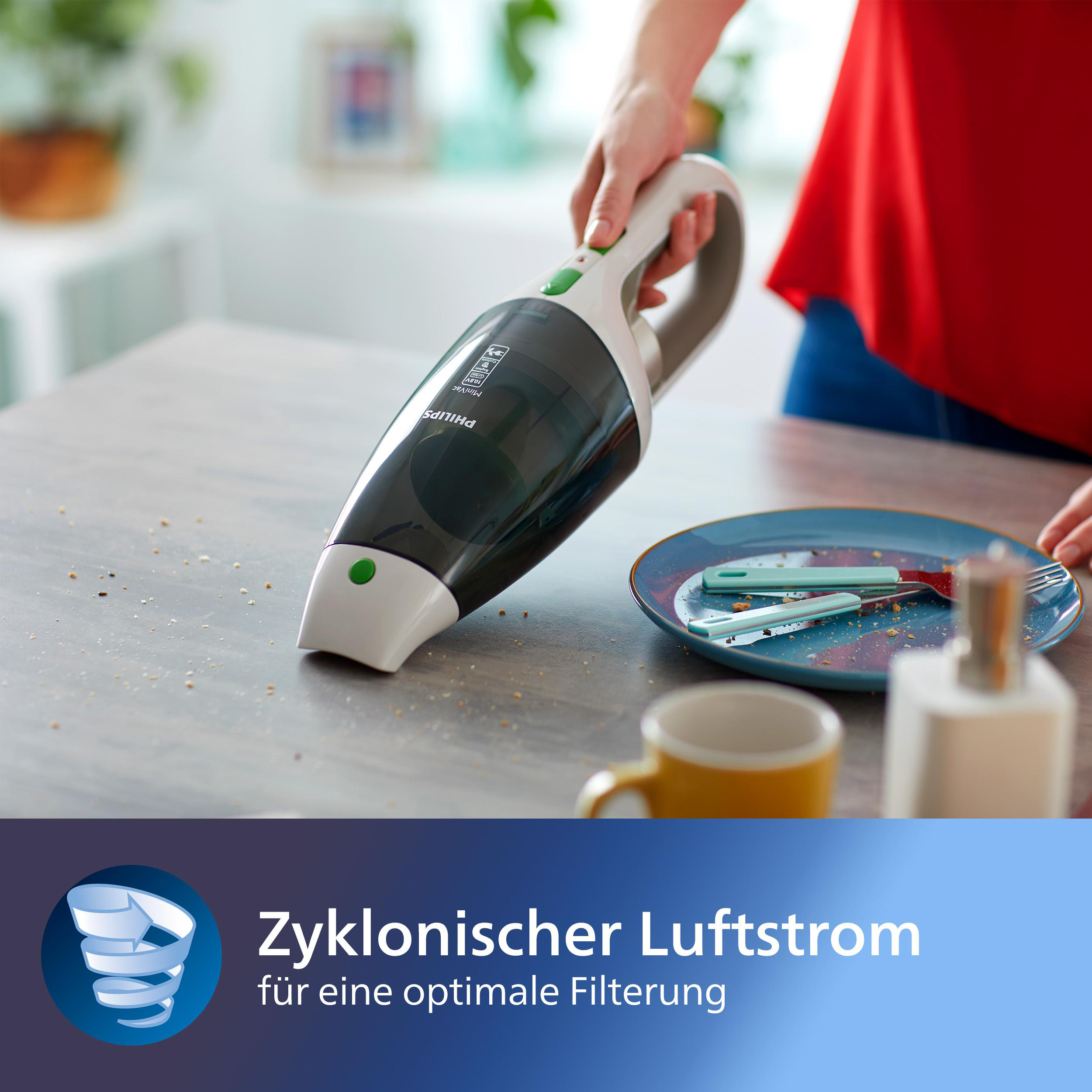 Ein Handstaubsauger reinigt Krümel auf einem Tisch. In der Nähe befinden sich ein Teller mit Besteck und eine Tasse. Im Hintergrund ein rotes Hemd.