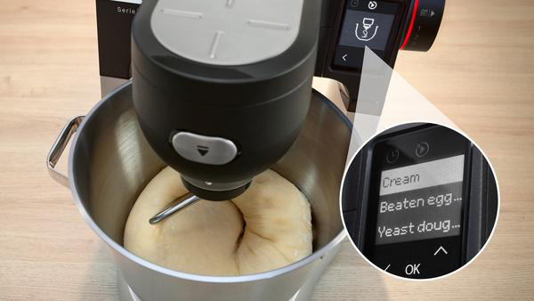 Ein Mixer mischt Teig. Ein Bildschirm zeigt Optionen. Der Hintergrund ist ein Tisch.