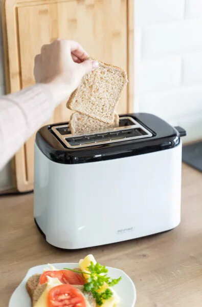 Osoba wkłada chleb do białego tostera. Talerz z jedzeniem na pierwszym planie.