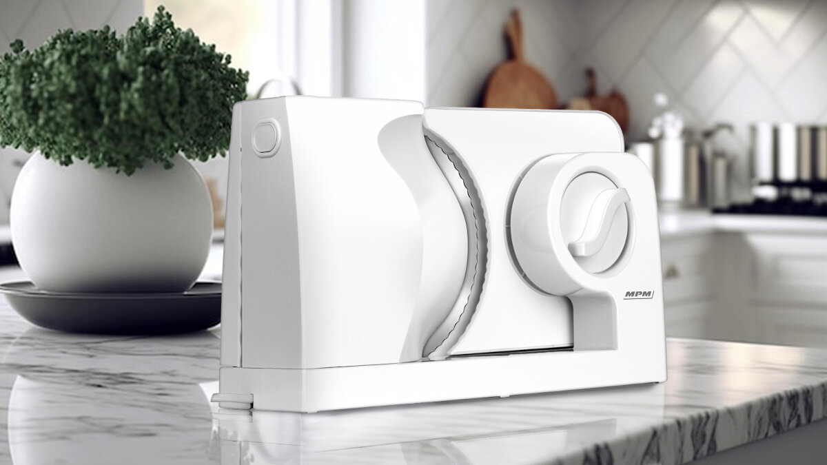 Biała krajalnica do żywności na marmurowym blacie, z rośliną i talerzem po lewej stronie. W tle kuchnia.