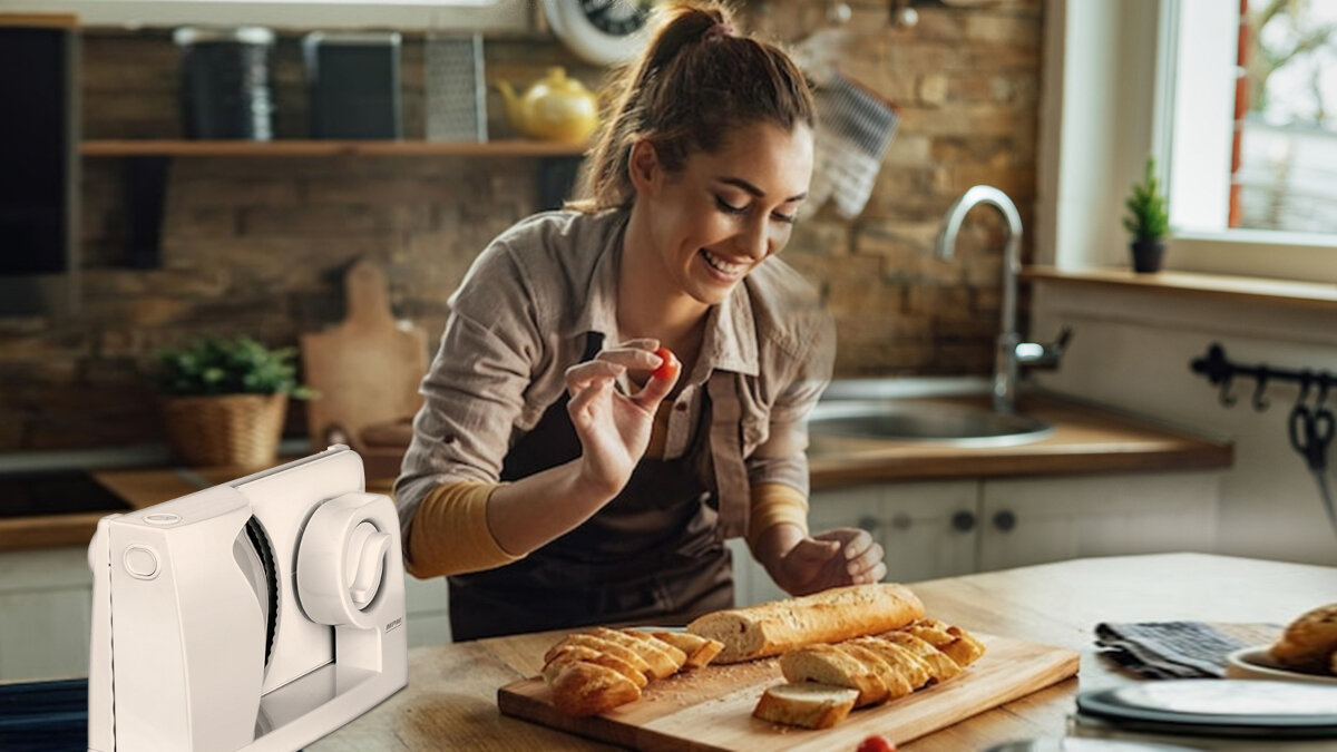 Kobieta uśmiecha się w kuchni, trzymając pomidora. Po lewej krajalnica do chleba, w środku pokrojony chleb.