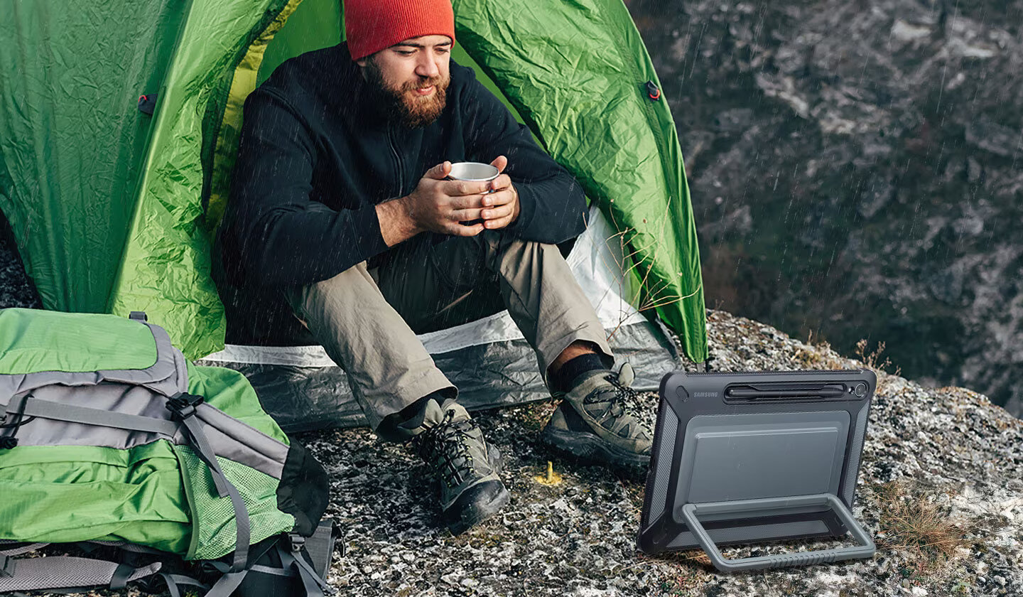 Mężczyzna w namiocie, trzymający kubek, w czapce i kurtce. Tablet na ziemi. Efekt deszczu.