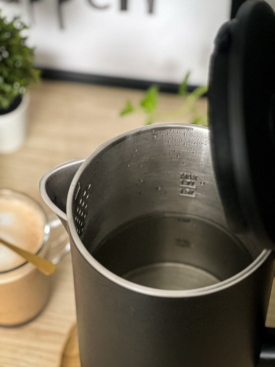 Czarny czajnik elektryczny, częściowo otwarty. W środku jest woda. W tle filiżanka kawy.