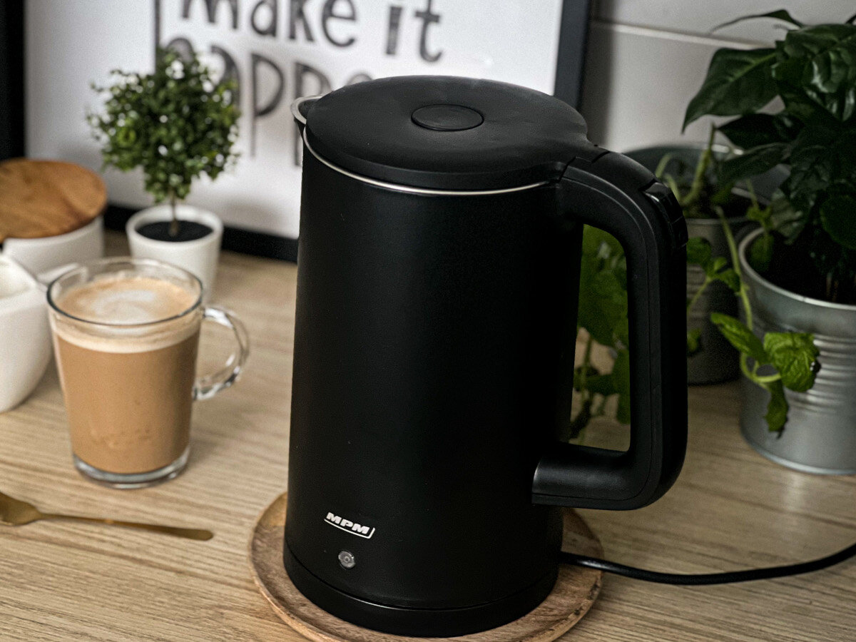 Czajnik elektryczny na drewnianej podstawce, obok filiżanki kawy i roślin.