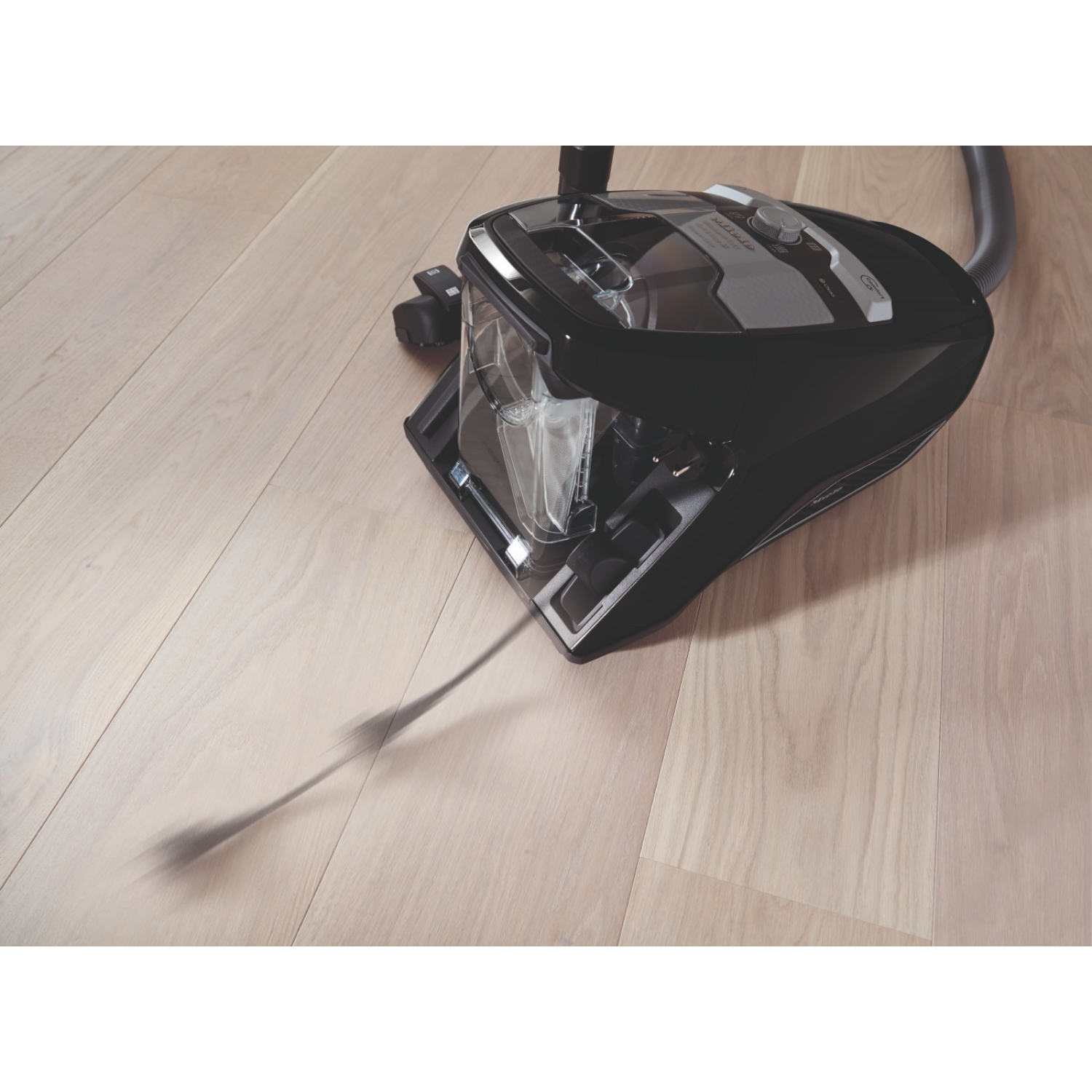 A black vacuum cleaner on a wooden floor. The dust container is transparent. The hose and accessories are visible.