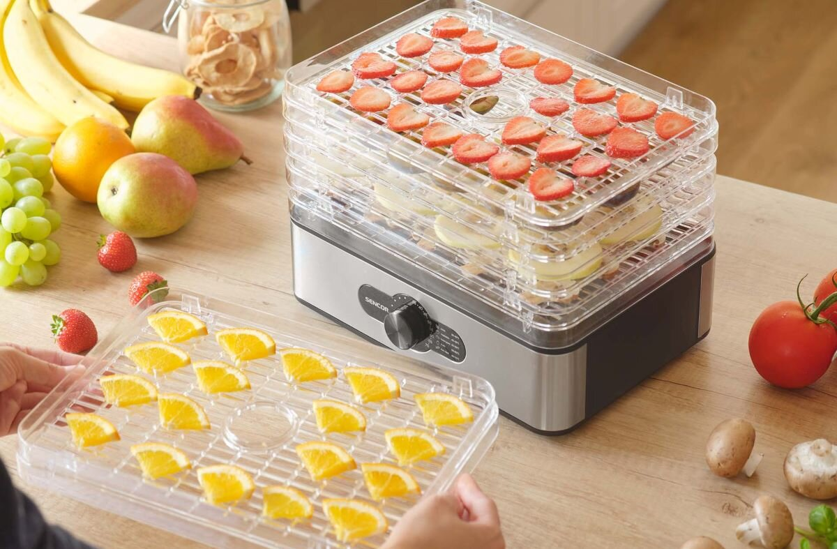 Suszarka do żywności ze stosami tacek z truskawkami i plasterkami pomarańczy. Owoce i warzywa obok.