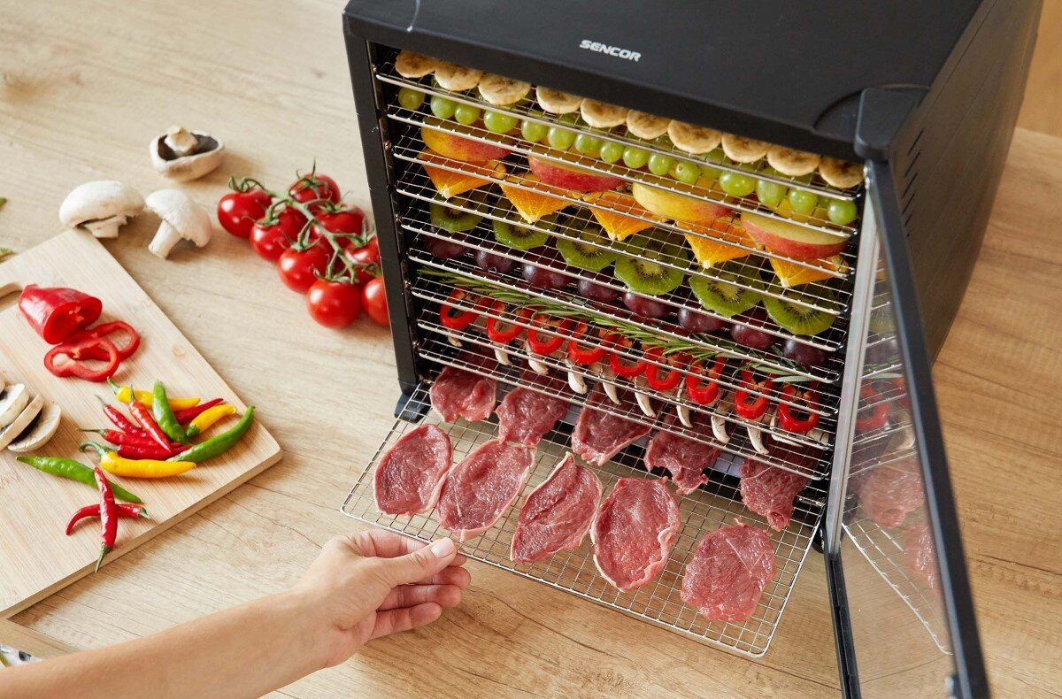 Suszarka do żywności z tacami owoców, mięsa i warzyw. Ręka sięga po tacę.
