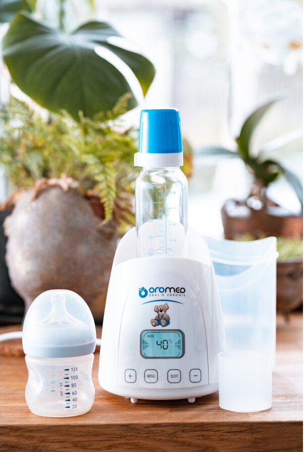 Podgrzewacz do butelek dla niemowląt z butelką, kubkiem i butelką. Drewniana powierzchnia, rośliny w tle.