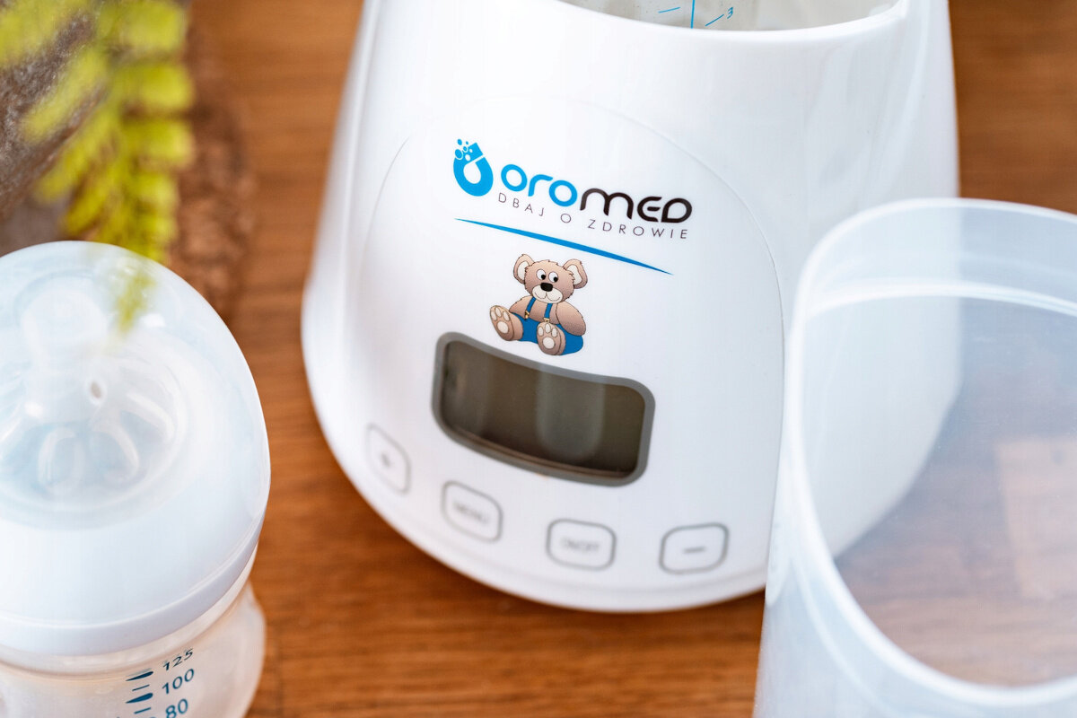 Biały podgrzewacz do butelek dla niemowląt. Butelki, pokrywka i logo misia. Drewniany stół.