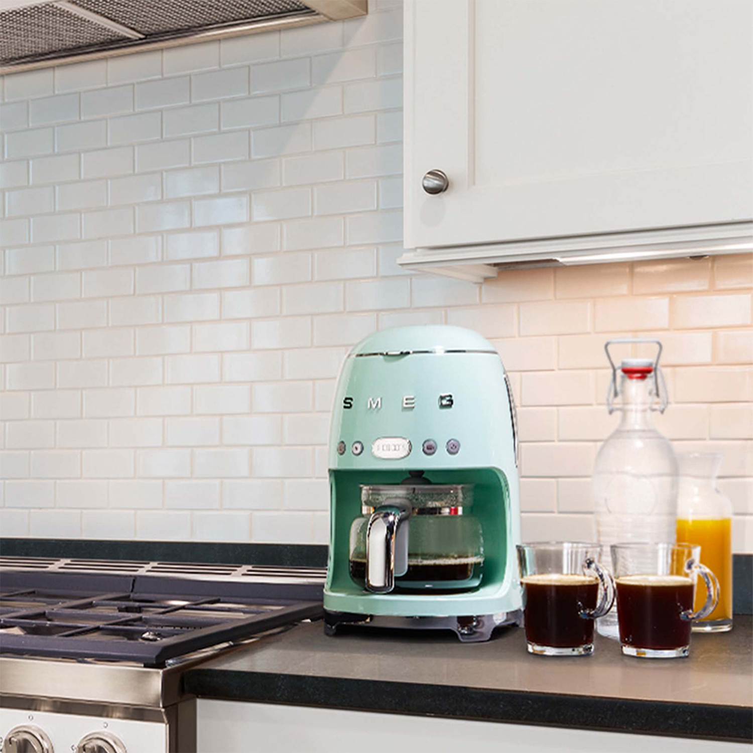 Caffettiera SMEG verde menta con caraffa in vetro, due tazze di caffè e una bottiglia d'acqua.