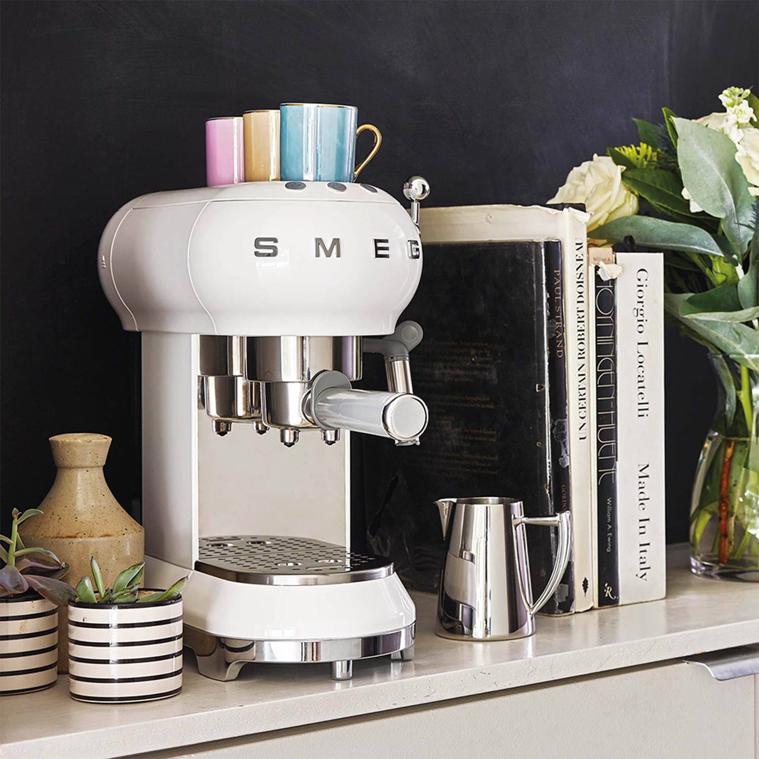 Una macchina per caffè espresso SMEG bianca su un piano di lavoro bianco con altri oggetti come libri, tazze e piante.