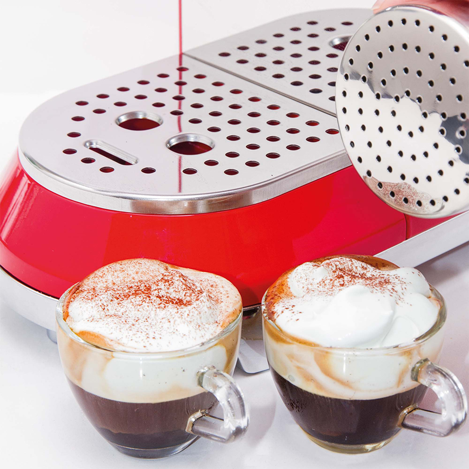 Due tazze di cappuccino vengono cosparse di cacao in polvere. Una macchina per espresso rossa è visibile.