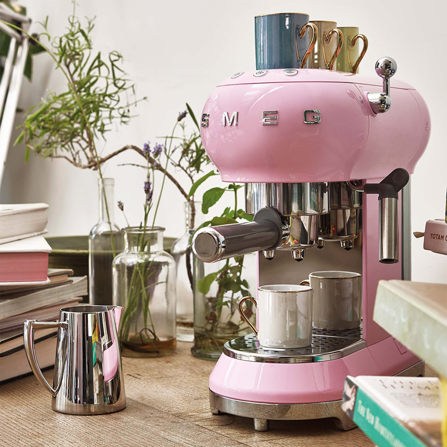 Macchina per espresso Smeg rosa su un tavolo di legno, con tazze e piante intorno.