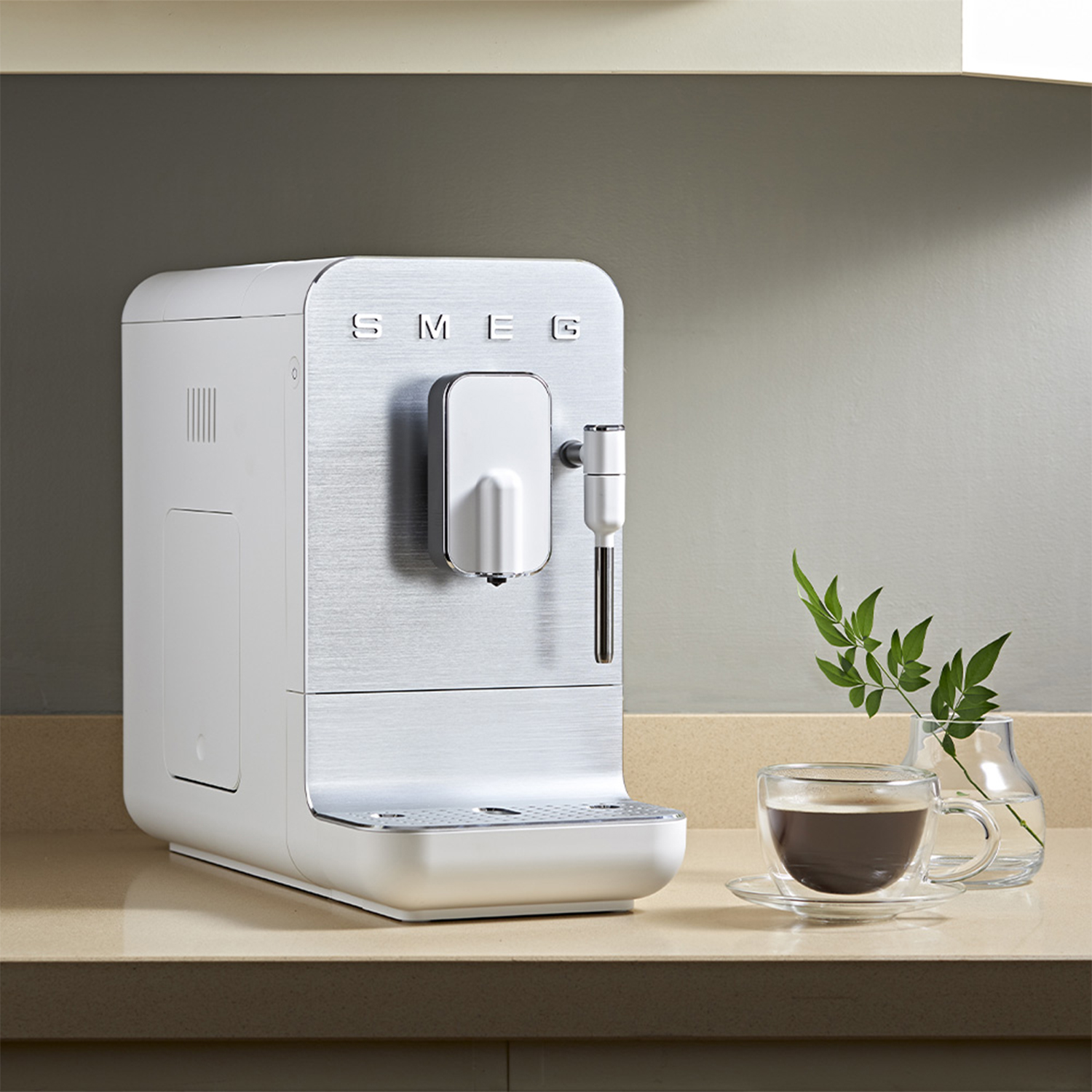 Macchina da caffè SMEG bianca con una tazza di vetro e una piccola pianta su una superficie beige.