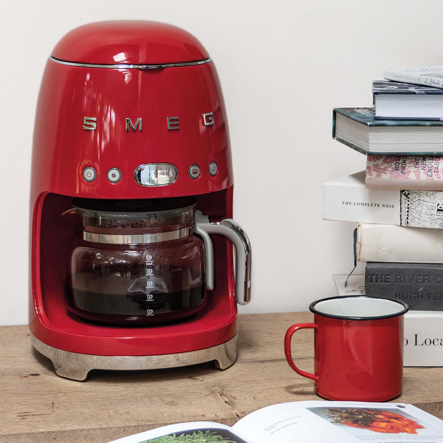 Caffettiera Smeg rossa con caraffa in vetro, tazza rossa e libri su una superficie in legno.