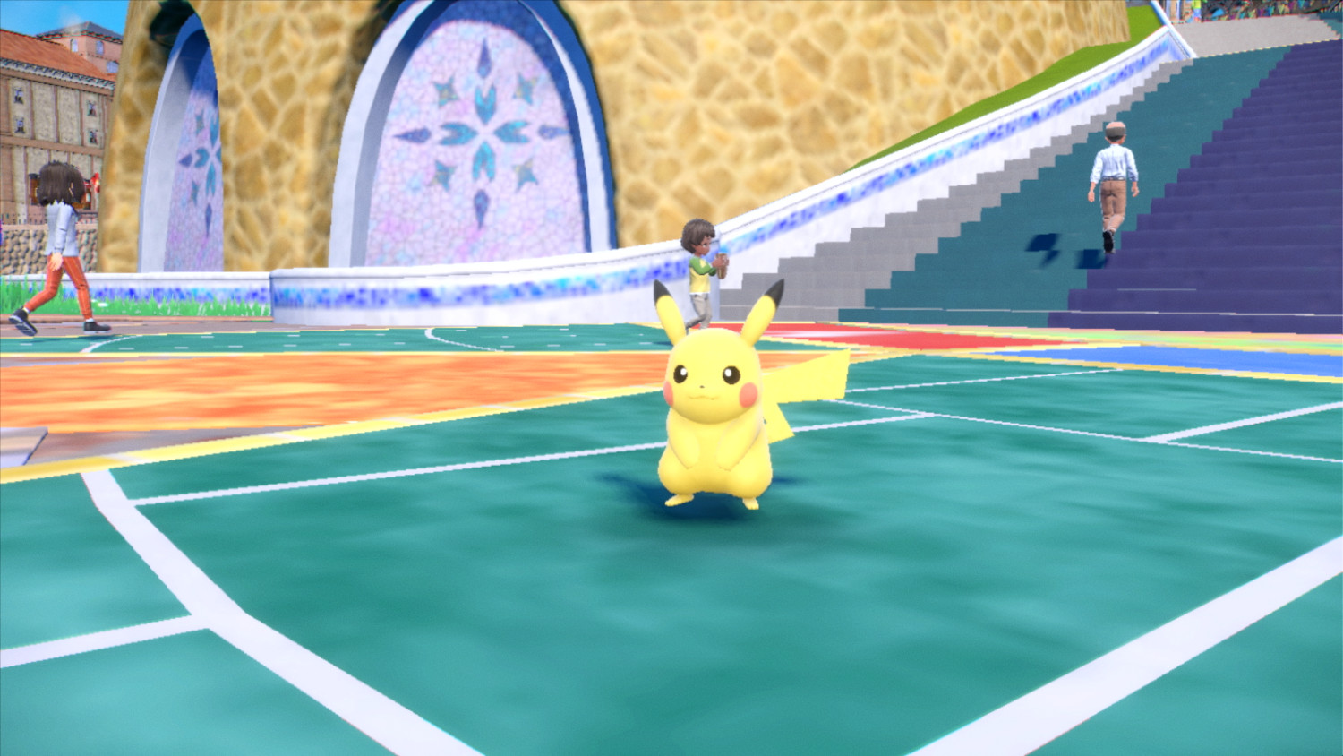 Pikachu staat op een groen en blauw veld, twee mensen lopen op de achtergrond.
