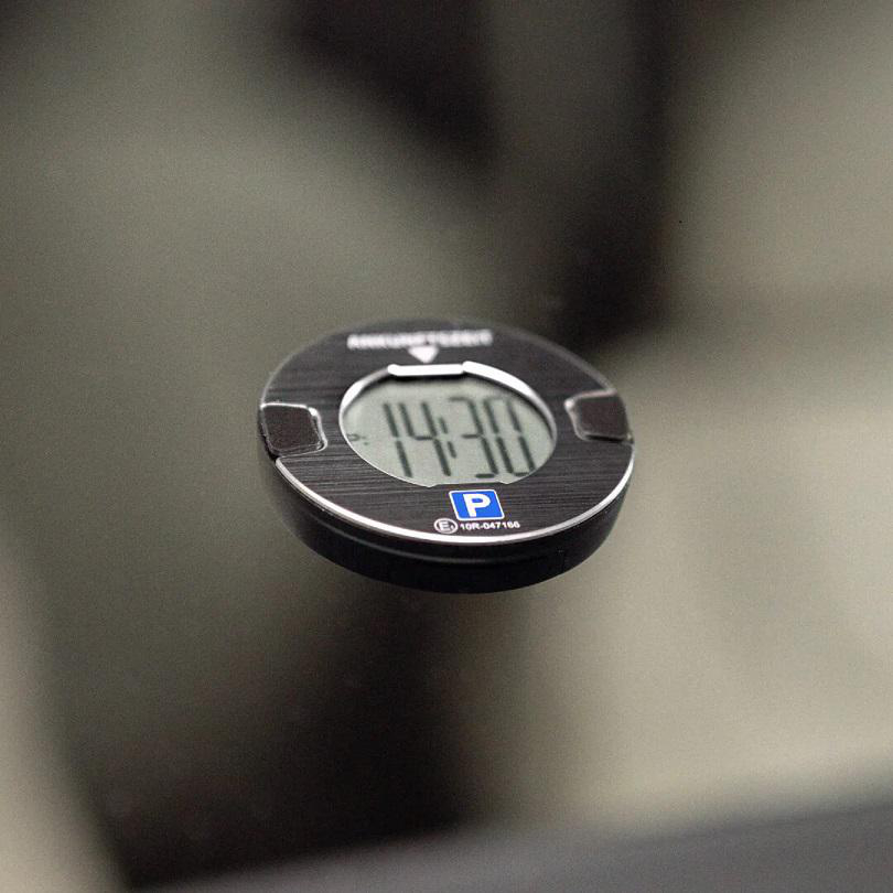 Eine schwarze Parkscheibe mit der Anzeige 14:30 und einem 'P' -Symbol auf einer reflektierenden Oberfläche.