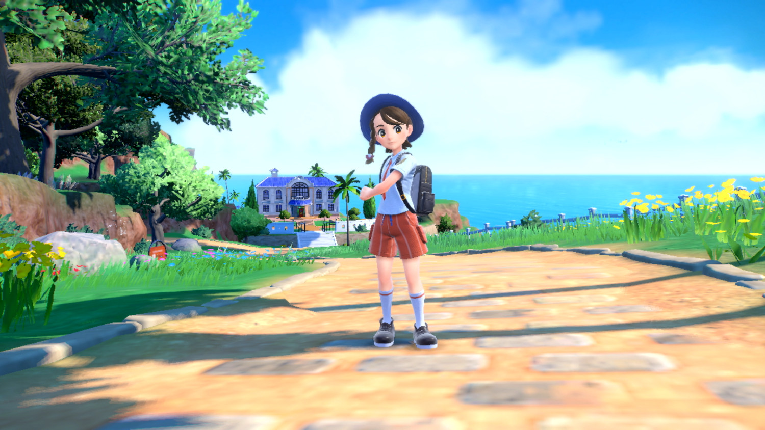 Een meisje met een blauwe hoed en schooluniform staat op een pad bij een strand met gebouwen.