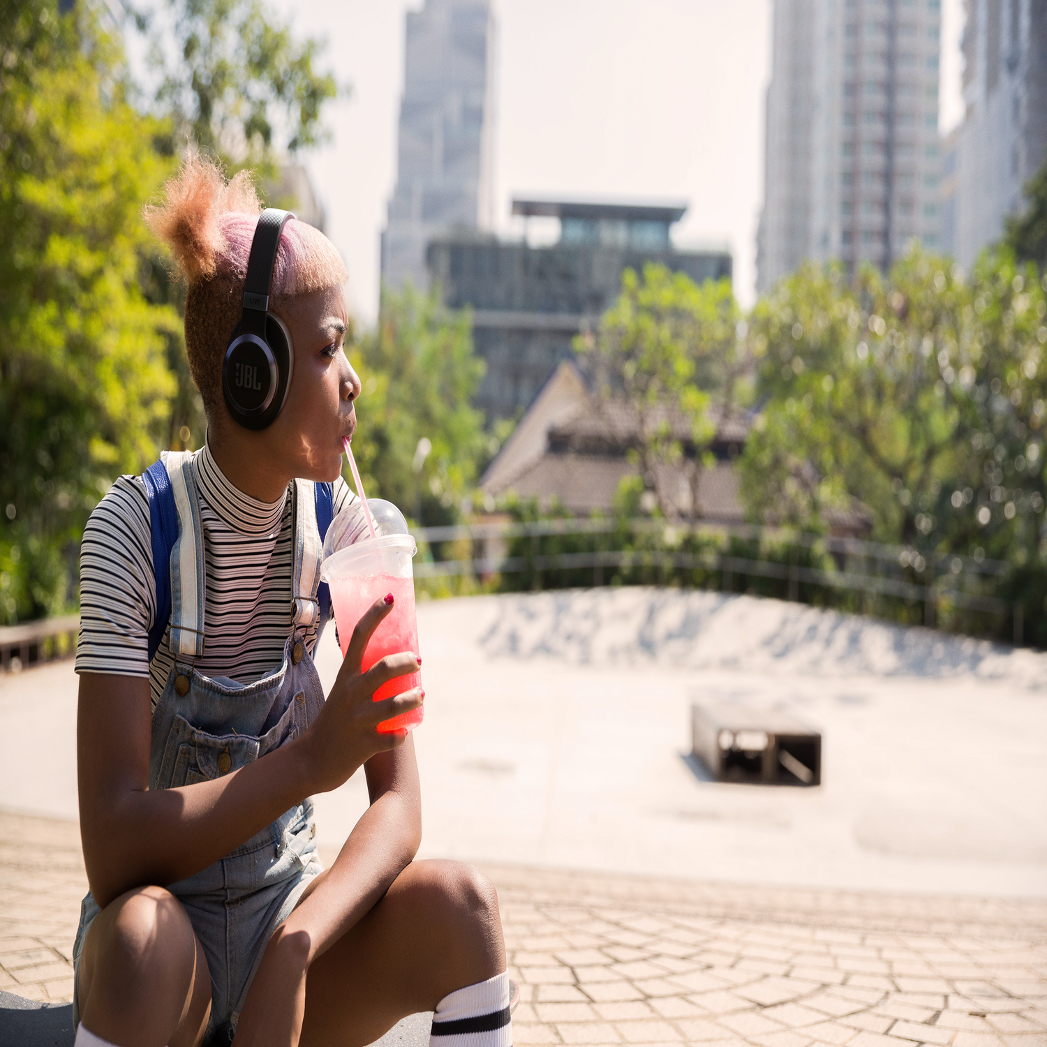Een jonge vrouw met roze haar en een koptelefoon drinkt uit een beker met een rietje, in een park.