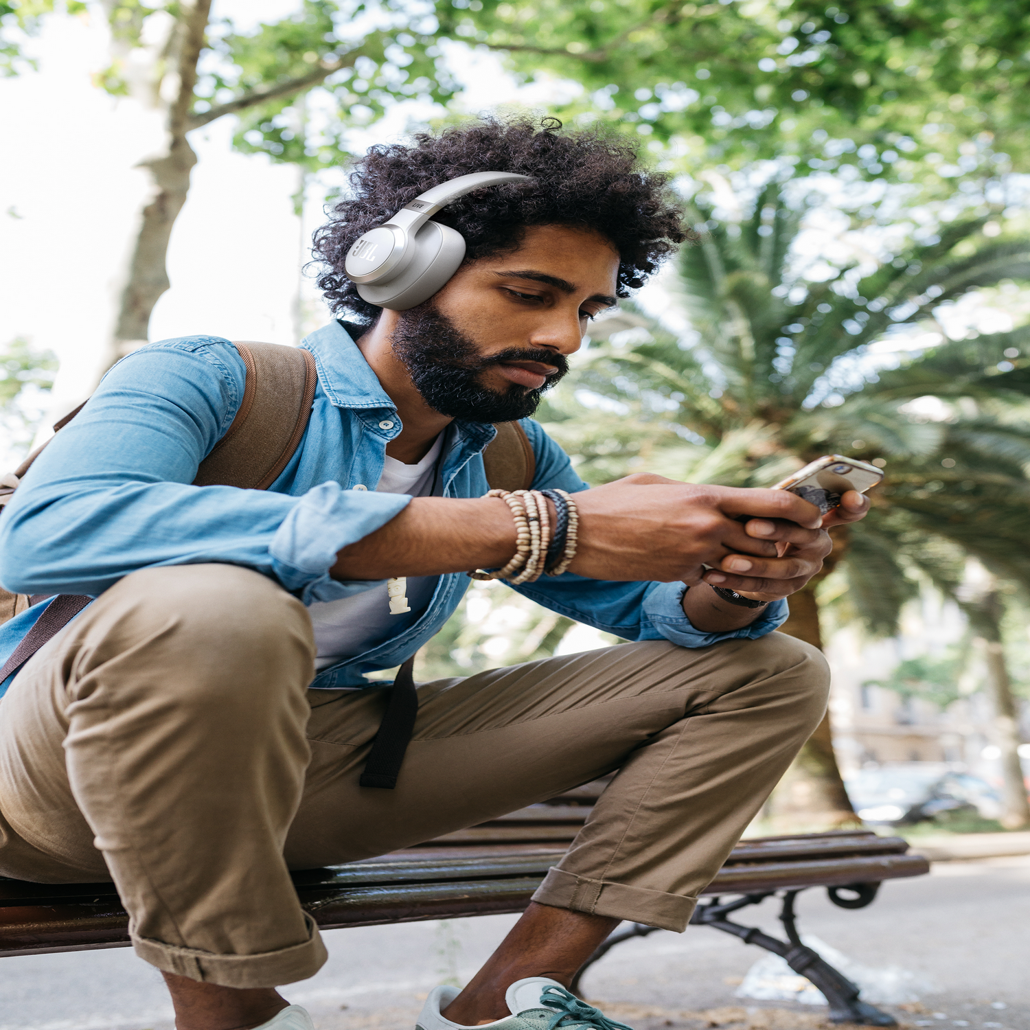 Ein Mann mit lockigem Haar hört Kopfhörer und schaut auf sein Handy. Er sitzt draußen auf einer Bank.