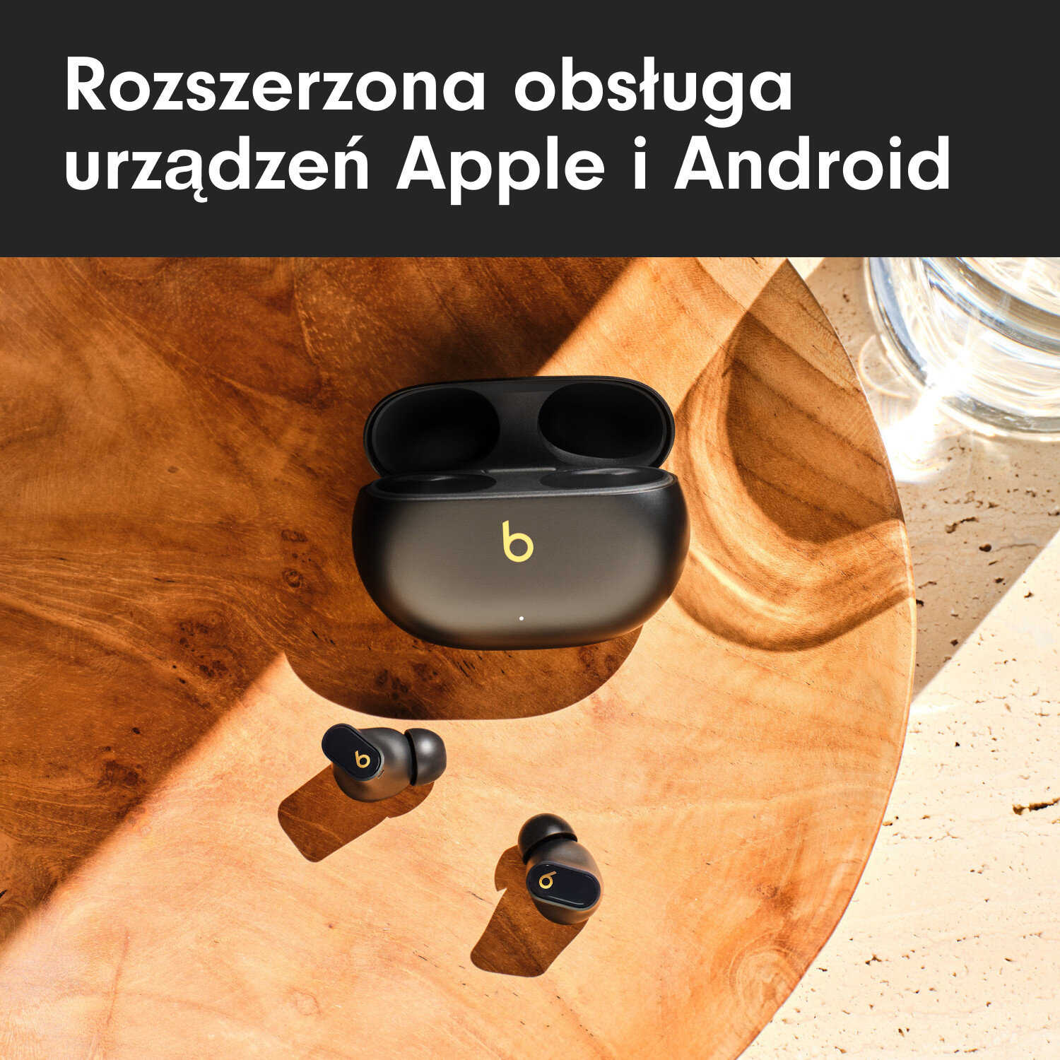 Czarne bezprzewodowe słuchawki w etui ładującym. Drewniana powierzchnia. Tekst w języku polskim u góry.