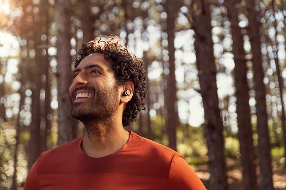 Mężczyzna w słuchawkach uśmiecha się, patrząc w górę, w leśnym otoczeniu. Drzewa w tle.