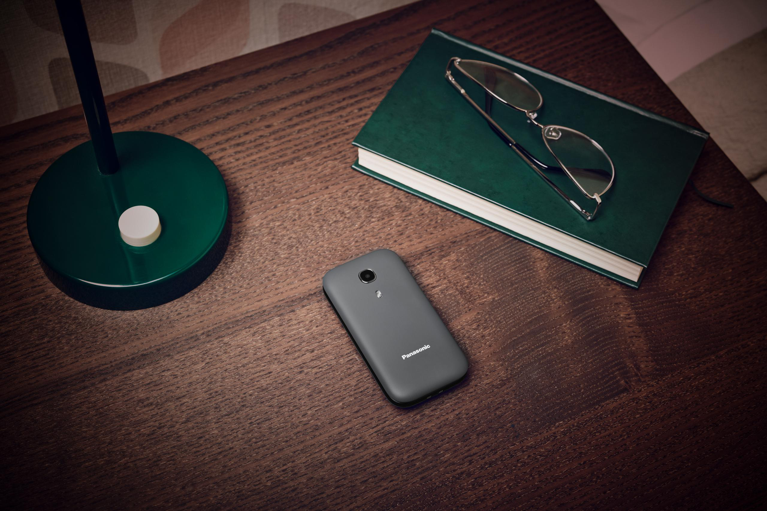 Graues Panasonic-Telefon auf einem Holztisch, mit Brille und Buch.