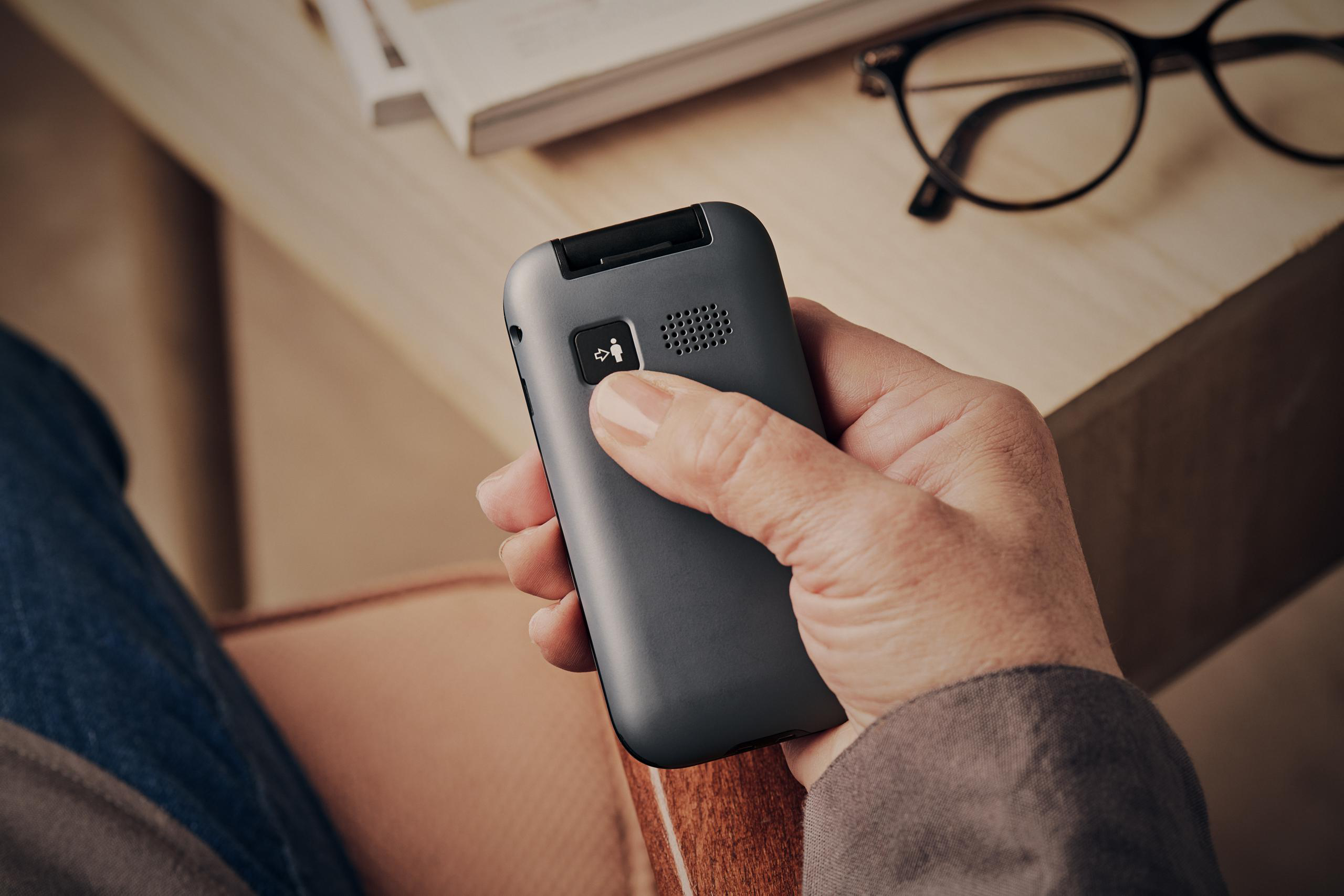 Hand, die ein graues Telefon hält. Im Hintergrund ein Tisch mit Brille und Buch.